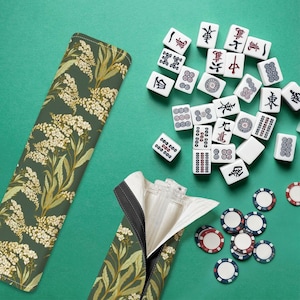 Puede incluir: Un estuche verde para fichas de Mahjong con un estampado floral, abierto para revelar un interior blanco. Las fichas de Mahjong y las fichas de juego están esparcidas sobre una superficie verde. Las fichas tienen caracteres y símbolos chinos.