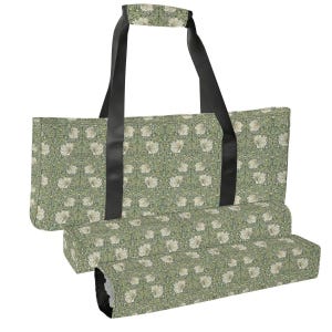 May include: A rectangular tote bag featuring a repeating floral pattern of white flowers and green foliage on a sage green background. The bag has two long black shoulder straps and a matching padded handle wrap with the same floral print.
