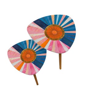May include: A set of two colorful, triangular side tables with a geometric design. The tables feature a pink, orange, blue, and white pattern with a gold circle in the center. The tables have wooden legs.