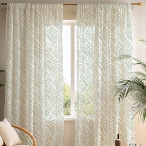 May include: Two light green and white patterned curtains with a leaf design hang in front of a window. The curtains are made of a lightweight fabric and are hanging from a wooden rod. A wicker chair and a potted plant are visible.