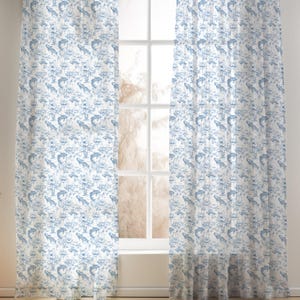 May include: Two sheer window curtains with a blue and white toile pattern. The pattern features aquatic life and floral elements. The curtains are hanging in front of a white window, allowing natural light to filter through.