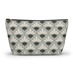 May include: A rectangular cosmetic bag with a repeating geometric pattern in shades of grey, black, and cream. The pattern features fan-like shapes and diamond shapes. The bag has a gold zip and a black zip pull.