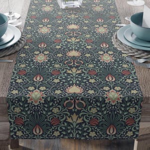 May include: A dark blue table runner with a repeating floral pattern in shades of green, red, and yellow. The runner is placed on a wooden table, with place settings visible on either side. The floral design is intricate and detailed.