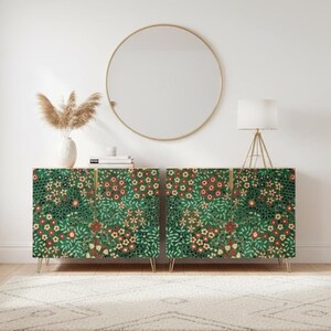 May include: Two matching green cabinets with floral patterns and gold legs. The cabinets are placed side by side, with a round gold mirror hanging above them. A white lamp and vase with dried plants are also visible.