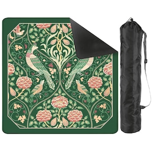 May include: A square outdoor blanket featuring a dark green background with a repeating pattern of birds, pink flowers, and cream-colored foliage. The blanket has a black underside and comes with a black cylindrical carrying bag with a shoulder strap.
