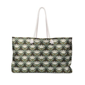 May include: Large tote bag with a repeating floral pattern in white, green, and brown on a dark background. The bag has white rope handles and is designed for carrying items. The bag is rectangular in shape.