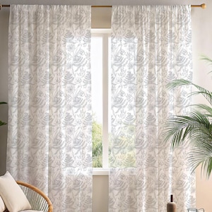 May include: Sheer white curtains with a gray botanical pattern. The curtains are hanging in front of a window, and the light filters through the fabric. The design features ferns and other plant elements, creating a natural aesthetic.