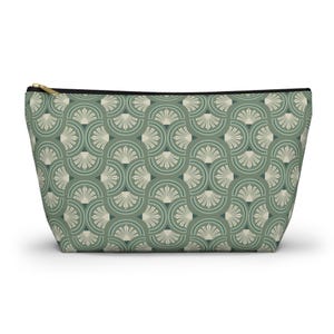 May include: A zipped cosmetic bag featuring a repeating fan-shaped pattern in shades of green and cream. The bag has a black zip and a gold-coloured pull. The bag is designed to hold makeup and other beauty products.