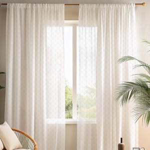 May include: Sheer white curtains hanging from a wooden rod, framing a bright window. The curtains have a subtle, repeating pattern and allow natural light to filter through. A wicker chair and a potted plant are visible in the corner.