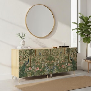 May include: A light wood cabinet with a botanical print featuring peacocks, trees, and flowers. The cabinet has gold-colored hairpin legs and a round gold mirror above it. A vase with greenery and a small tray are on top.