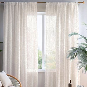 May include: Sheer, cream-colored curtains with a repeating white palm leaf pattern. The curtains hang in front of a window, allowing natural light to filter through. A wooden curtain rod is visible at the top. The curtains add a touch of tropical style to the room.