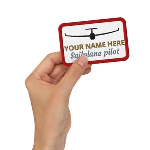 May include: A rectangular patch with a red border and white center. The patch features a black glider illustration above the text "YOUR NAME HERE" in gold and "Sailplane pilot" in gray.