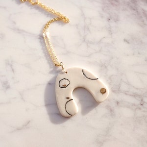 Op de afbeelding: Een witte keramische ketting met een gouden ketting. De hanger is gevormd als een boog met zwarte en gouden abstracte ontwerpen.