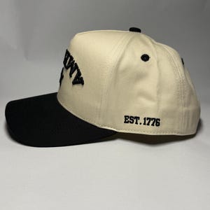 K&ouml;nnte beinhalten: Cremefarbene Baseballkappe mit schwarzem Schirm. Auf der Kappe ist das Wort "AMMY" in Schwarz auf der Vorderseite und "EST. 1776" auf der Seite gestickt. Die Kappe hat einen gebogenen Schirm und einen Knopf oben.