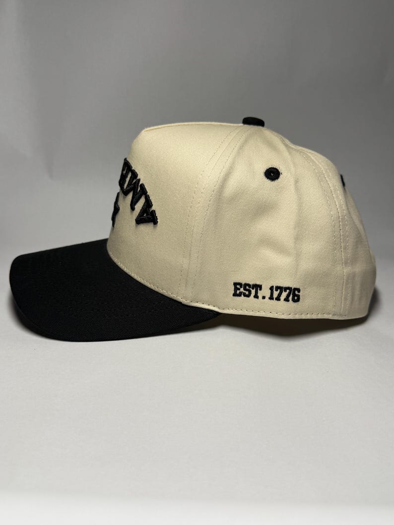 K&ouml;nnte beinhalten: Cremefarbene Baseballkappe mit schwarzem Schirm. Auf der Kappe ist das Wort "AMMY" in Schwarz auf der Vorderseite und "EST. 1776" auf der Seite gestickt. Die Kappe hat einen gebogenen Schirm und einen Knopf oben.