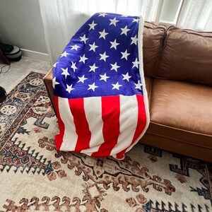 May include: A red, white, and blue American flag blanket draped over a brown leather couch. The blanket is made of soft, plush fabric and features a white star pattern on a blue background.