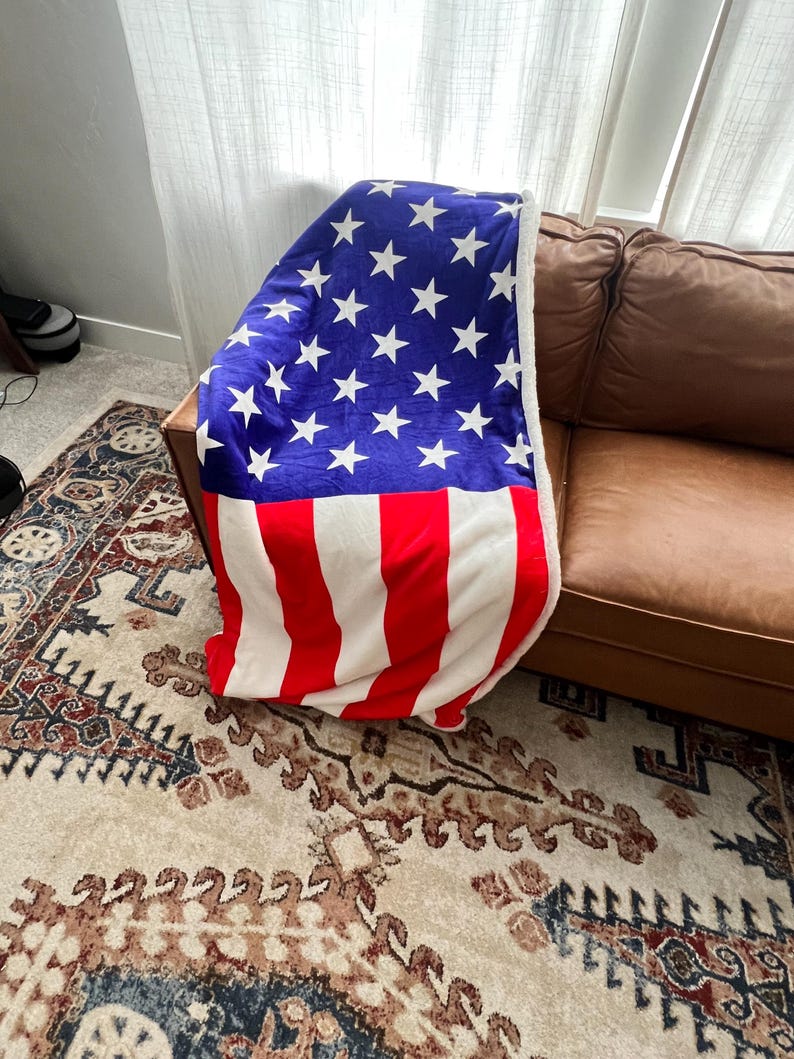 May include: A red, white, and blue American flag blanket draped over a brown leather couch. The blanket is made of soft, plush fabric and features a white star pattern on a blue background.