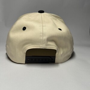 K&ouml;nnte beinhalten: Beige Baseball Cap mit schwarzem Schirm und schwarzem Knopf oben. Die Kappe hat einen Snapback-Verschluss f&uuml;r eine verstellbare Gr&ouml;&szlig;e. Die Kappe hat zwei kleine schwarze &Ouml;sen an der Seite.