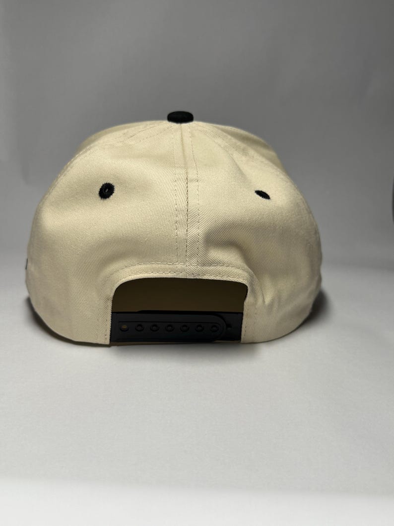 K&ouml;nnte beinhalten: Beige Baseball Cap mit schwarzem Schirm und schwarzem Knopf oben. Die Kappe hat einen Snapback-Verschluss f&uuml;r eine verstellbare Gr&ouml;&szlig;e. Die Kappe hat zwei kleine schwarze &Ouml;sen an der Seite.