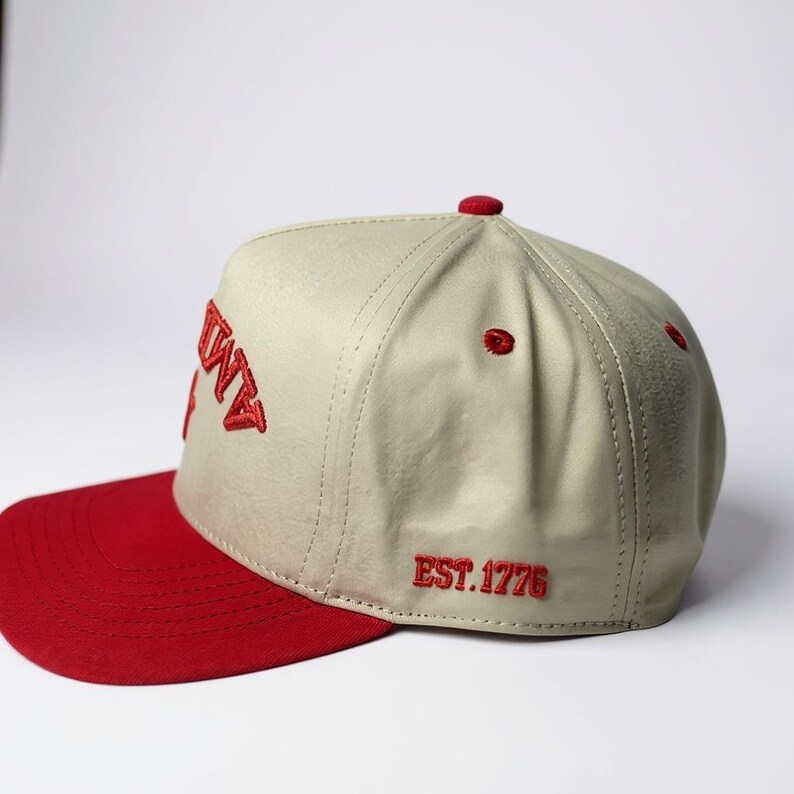 Peut inclure: Casquette de baseball beige et rouge avec le mot "AMITY" brod&eacute; en rouge sur le devant. La visi&egrave;re est rouge et la casquette porte la mention "EST. 1776" brod&eacute;e sur le c&ocirc;t&eacute;. La casquette a une visi&egrave;re incurv&eacute;e et un bouton sur le dessus.