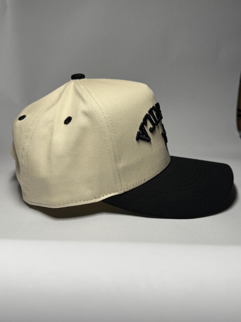 K&ouml;nnte beinhalten: Beige-schwarze Baseballkappe mit dem Wort "RICA" in Schwarz auf der Vorderseite gestickt. Die Kappe hat eine strukturierte Krone und einen gebogenen Schirm. Hergestellt aus strapazierf&auml;higem Stoff.