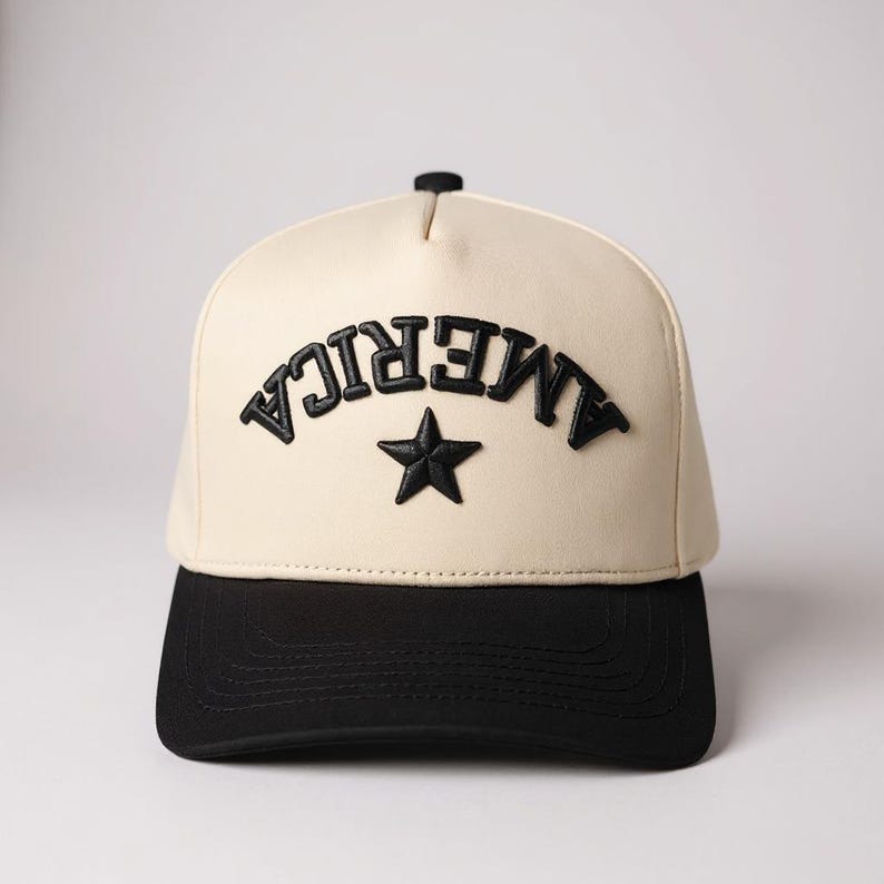 K&ouml;nnte beinhalten: Cremefarbene Baseballkappe mit schwarzem Schirm. Das Wort "AMERICA" ist in erhabenen schwarzen Buchstaben auf der Vorderseite aufgebogen, darunter ein schwarzer Stern. Ein stilvolles Accessoire.