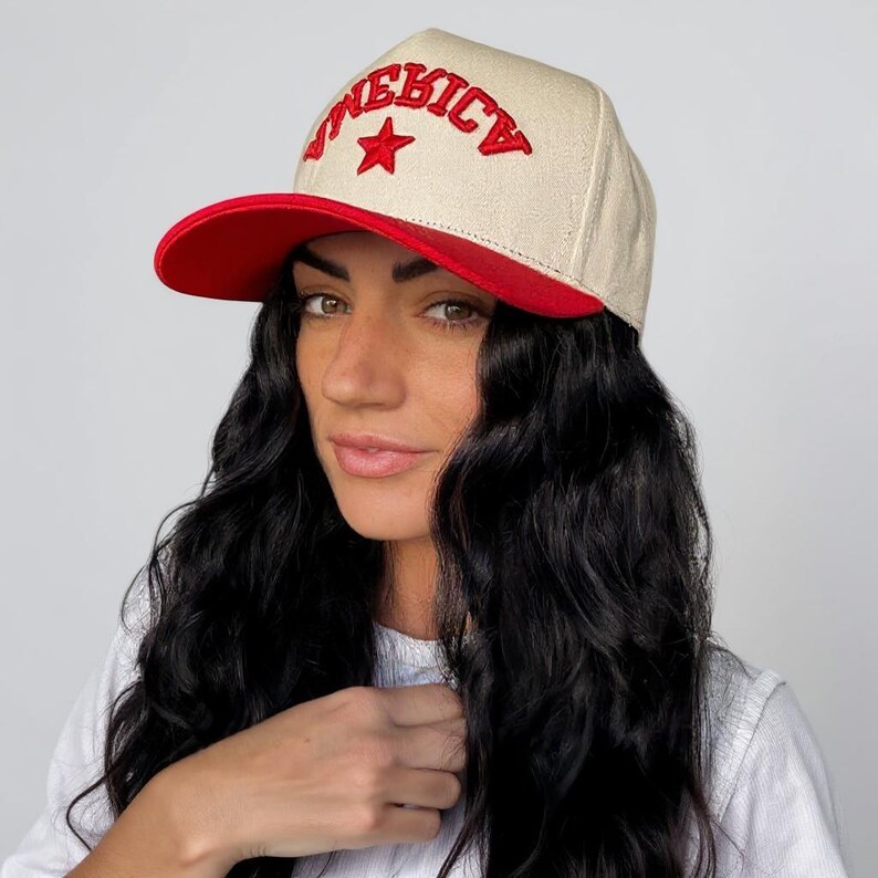 Peut inclure: Casquette de baseball beige avec une visi&egrave;re rouge et le mot "AMERICA" brod&eacute; en rouge, ainsi qu'une &eacute;toile rouge. La casquette est port&eacute;e par une personne aux longs cheveux noirs.