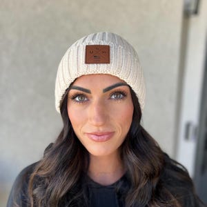 Pode incluir: Um gorro de malha creme com um remendo de couro marrom que diz "USA" com uma pequena bandeira americana.