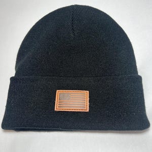 Puede incluir: Gorro negro con un parche de cuero marrón con un diseño de la bandera estadounidense. El gorro está hecho de un material de punto suave y tiene un borde doblado. El parche de la bandera es rectangular y está cosido en la parte delantera.