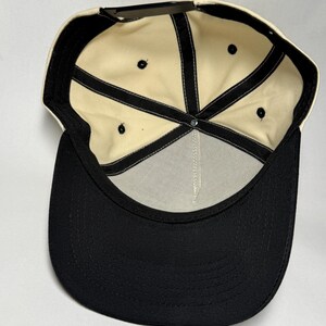 K&ouml;nnte beinhalten: Eine Baseballkappe in Schwarz und Creme. Die Kappe hat einen schwarzen Schirm und eine cremefarbene Krone. Die Innenseite hat ein cremefarbenes Futter mit schwarzen N&auml;hten und einen schwarzen, verstellbaren Riemen.