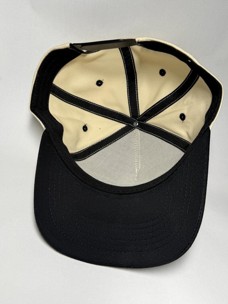 K&ouml;nnte beinhalten: Eine Baseballkappe in Schwarz und Creme. Die Kappe hat einen schwarzen Schirm und eine cremefarbene Krone. Die Innenseite hat ein cremefarbenes Futter mit schwarzen N&auml;hten und einen schwarzen, verstellbaren Riemen.