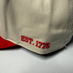 Peut inclure: Casquette de baseball avec une visi&egrave;re rouge et le texte "EST. 1776" brod&eacute; en rouge sur le c&ocirc;t&eacute;. La couronne de la casquette est de couleur beige clair.