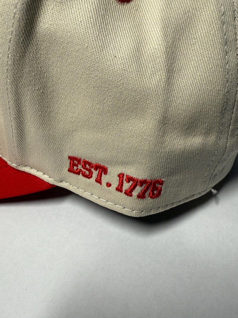 Peut inclure: Casquette de baseball avec une visi&egrave;re rouge et le texte "EST. 1776" brod&eacute; en rouge sur le c&ocirc;t&eacute;. La couronne de la casquette est de couleur beige clair.