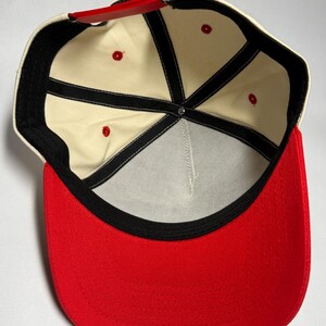 Peut inclure: Casquette de baseball avec une visi&egrave;re rouge et une couronne cr&egrave;me. L'int&eacute;rieur pr&eacute;sente une doublure cr&egrave;me avec des coutures noires et des &oelig;illets rouges. La sangle r&eacute;glable est rouge.