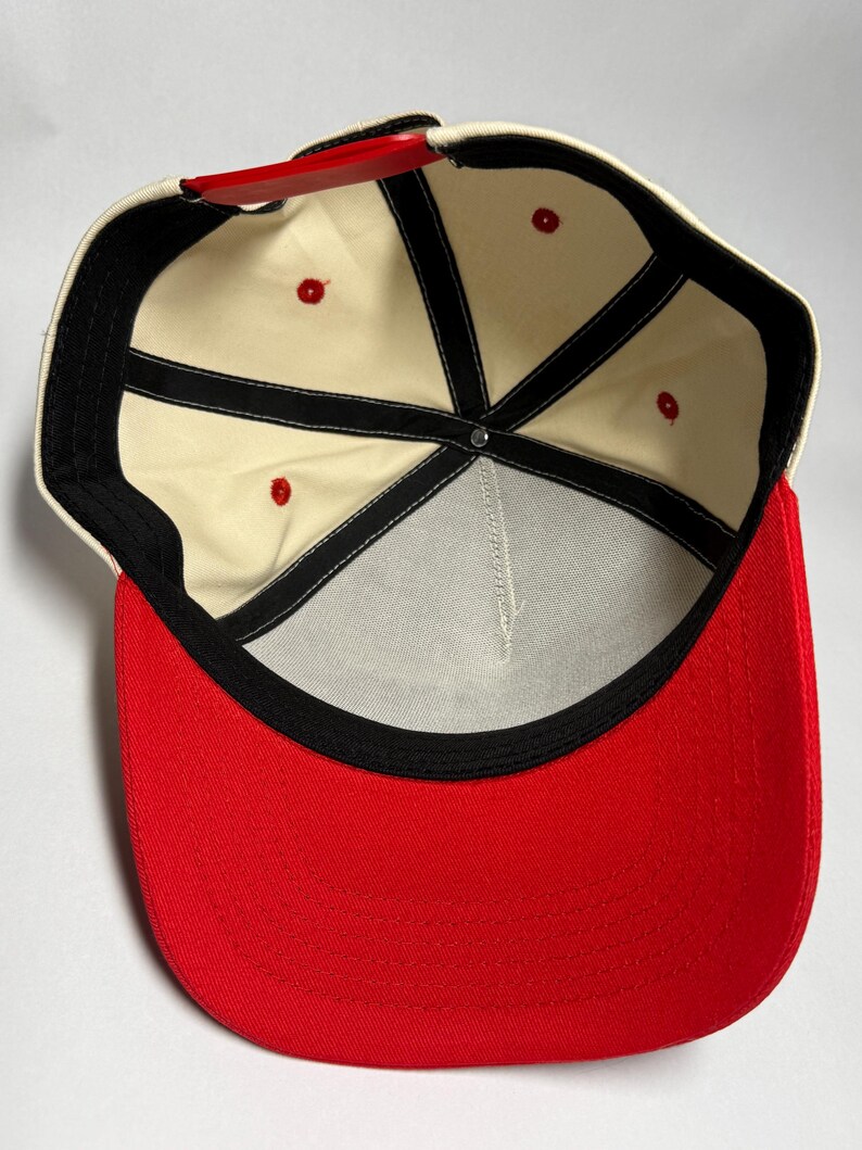 Peut inclure: Casquette de baseball avec une visi&egrave;re rouge et une couronne cr&egrave;me. L'int&eacute;rieur pr&eacute;sente une doublure cr&egrave;me avec des coutures noires et des &oelig;illets rouges. La sangle r&eacute;glable est rouge.
