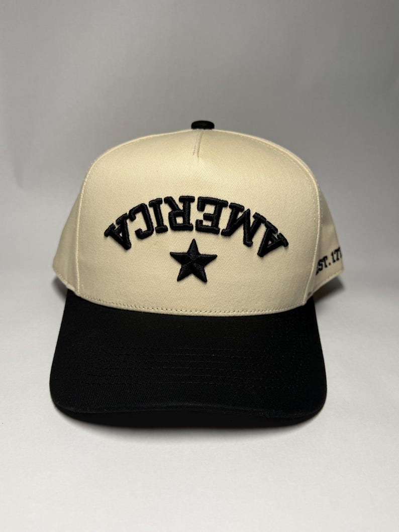 K&ouml;nnte beinhalten: Cremefarbene Baseballkappe mit schwarzem Schirm. Auf der Vorderseite ist das Wort "AMERICA" in Schwarz gestickt, zusammen mit einem schwarzen Stern. Der Text "ST. 17" befindet sich an der Seite.