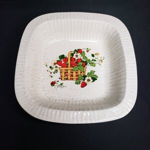 May include: Cream-colored, square serving dish with a ribbed edge and a speckled finish. The center features a basket of red strawberries with green leaves and white flowers. The text "Crispy Sims" is below the basket.