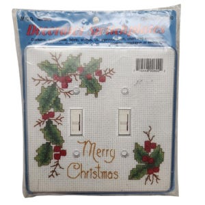 May include: A double light switch plate cover kit in original plastic packaging. The white plate features a cross-stitch pattern of green holly leaves and red berries with the text Merry Christmas in gold script. Includes two switch cutouts.