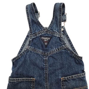May include: Dark blue denim overalls with adjustable straps and pockets. The overalls feature the "OshKosh B'gosh" logo on the bib. The garment has a classic design with visible stitching and metal hardware.