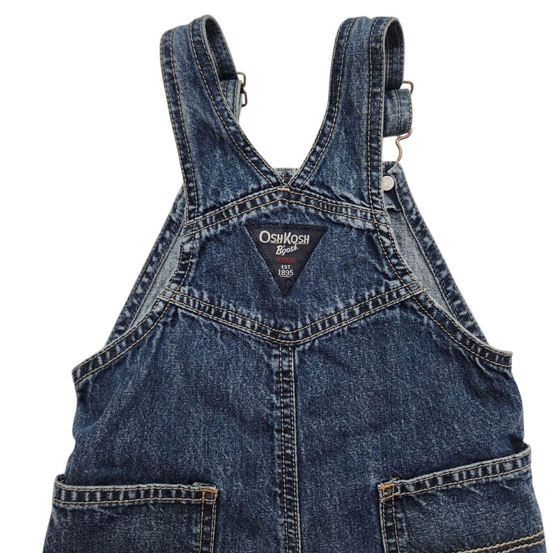 May include: Dark blue denim overalls with adjustable straps and pockets. The overalls feature the "OshKosh B'gosh" logo on the bib. The garment has a classic design with visible stitching and metal hardware.