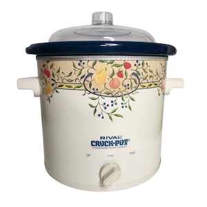 May include: A Rival Crock-Pot stoneware slow cooker with a cream-colored base, a dark blue rim, and a clear glass lid. The exterior features a decorative border of painted fruits and vines. A control knob is centered below the text RIVAL CROCK-POT.