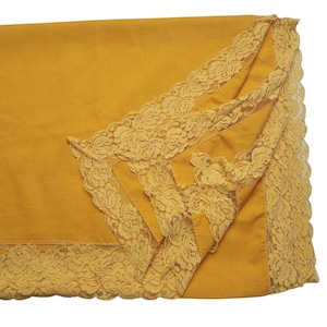 Puede incluir: Mantel mostaza con un delicado ribete de encaje. El ribete de encaje es de un tono amarillo más claro y está intrincadamente diseñado con patrones florales. El mantel está doblado, mostrando el detalle del encaje en los bordes.