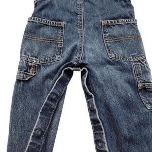 May include: Close-up of blue denim overalls with multiple pockets. The overalls have a classic design with reinforced stitching and a faded wash. The pockets are a medium size and are located on the back and sides of the overalls.