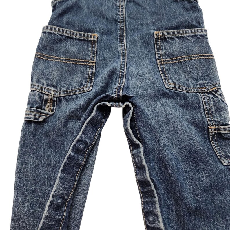 May include: Close-up of blue denim overalls with multiple pockets. The overalls have a classic design with reinforced stitching and a faded wash. The pockets are a medium size and are located on the back and sides of the overalls.