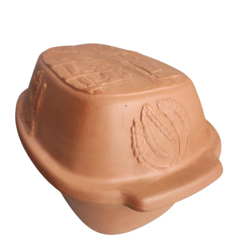 May include: A terracotta clay baker with a lid, featuring embossed wheat and farm scene designs. The oval-shaped baker is a warm, earthy brown color, ideal for baking bread or casseroles. The design is rustic and functional.