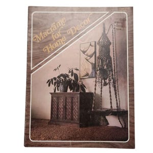 May include: A vintage book cover titled "Macrame for Home Decor" with a brown wooden frame. The image inside shows a macrame hanging chair, a wooden cabinet, and a potted plant. The background is a neutral color.