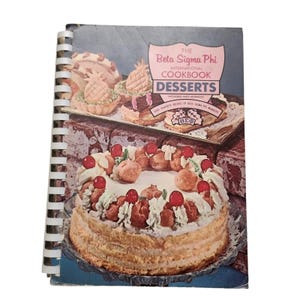 May include: A vintage cookbook titled "The Beta Sigma Phi International Cookbook Desserts." The cover shows a cake with cherries and cream puffs, and other desserts. The cookbook is spiral-bound.