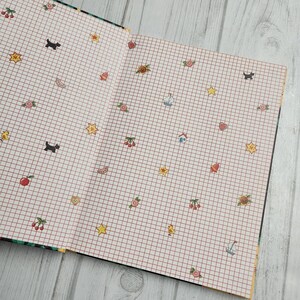 May include: An open notebook with a white grid pattern and red lines. The notebook is decorated with various small illustrations, including stars, hearts, cherries, sailboats, houses, and a sunflower.