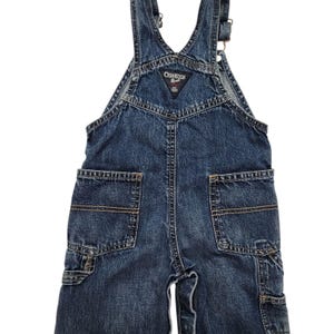 May include: Blue denim overalls with the OshKosh B'gosh logo on the bib. The overalls feature two front pockets, two back pockets, and adjustable straps. The denim is a medium wash, and the garment is designed for a child.