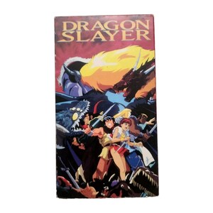 以下が含まれることがあります： 金色の文字で「DRAGON SLAYER」と書かれた長方形のVHSカバー。アニメ調のキャラクターたちが剣を構え、黄色い目をした暗い怪物や、炎のような色の大きなドラゴンの頭部を背景に描かれたイラストが特徴です。